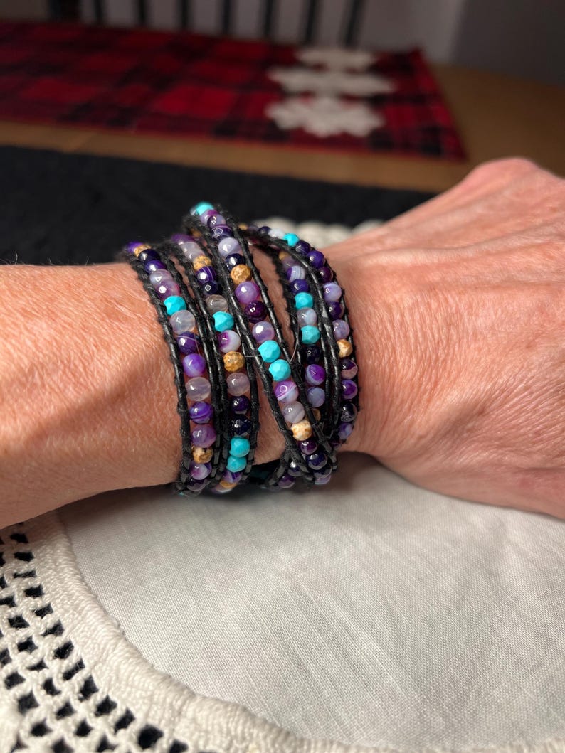 May include: Wrap bracelet featuring a multi-strand design with purple, turquoise, and tan beads. The bracelet is strung on black cord and wraps multiple times around the wrist. The beads are various sizes and shapes.