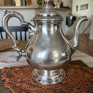 May include: A silver-colored teapot with a curved handle and spout, resting on a decorative base. The teapot has a lid with a pointed finial and is placed on a brown, open-weave doily.
