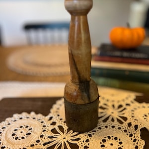 May include: A vintage wooden muddler with a light brown, ridged top and a dark brown handle. The muddler sits on a cream-colored, crocheted doily. An orange pumpkin and books are in the background.