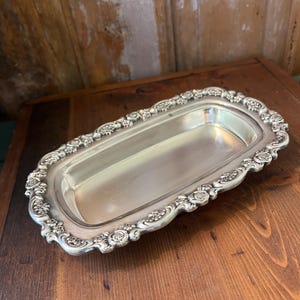 May include: A rectangular silver-toned metal tray featuring a smooth, recessed center and a raised, ornate border decorated with a repeating pattern of sculpted roses and small floral clusters along the entire outer edge.