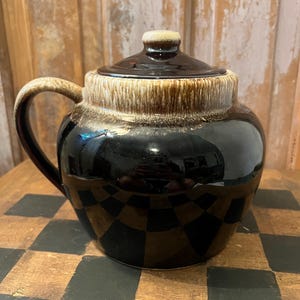 May include: A vintage ceramic jar with a lid. The jar is a glossy black with a tan and brown rim and lid. It has a curved handle and a small knob on top of the lid. The jar is sitting on a checkered surface.