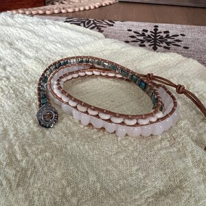 May include: Wrap bracelet featuring a combination of light pink, blue-green, and clear beads. The bracelet has a brown leather strap and a silver button clasp. The beads are arranged in multiple strands, creating a layered effect. The bracelet is displayed on a cream-colored fabric.