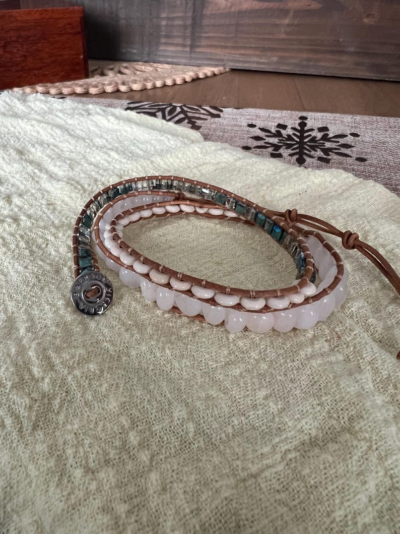May include: Wrap bracelet featuring a combination of light pink, blue-green, and clear beads. The bracelet has a brown leather strap and a silver button clasp. The beads are arranged in multiple strands, creating a layered effect. The bracelet is displayed on a cream-colored fabric.