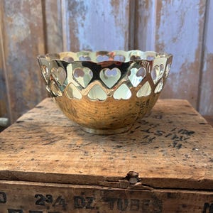 May include: A gold-colored bowl with heart-shaped cutouts around the rim. The bowl sits on a wooden crate with faded text. The bowl has a shiny, reflective surface and a rounded shape. The heart cutouts add a decorative touch to the bowl's design.
