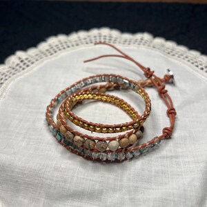 Może przedstawiać: Wielopasmowa bransoletka typu wrap z złotymi, brązowymi i przezroczystymi koralikami. Bransoletka, wykonana z brązowej sk&oacute;ry, owija się kilka razy wok&oacute;ł nadgarstka. Koraliki mają zr&oacute;żnicowane tekstury i kolory. Dodatek modowy.