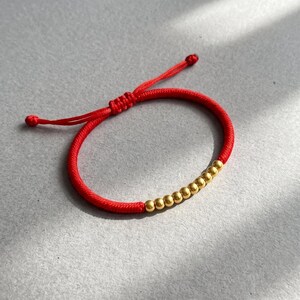 May include: A red cord bracelet featuring a series of ten small, round, metallic gold-coloured beads arranged in a line. The bracelet has an adjustable sliding knot closure with two loose cord ends extending from the central knot mechanism.