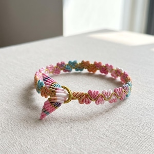 Può includere: Un braccialetto intrecciato caratterizzato da un motivo ripetitivo di piccoli nodi a forma di cuore in fili color pastello rosa, blu e arancione. Un anello metallico dorato collega le estremità, con un'estensione decorativa a coda in fili abbinati.
