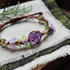 May include: A brown cord bracelet featuring a central raw amethyst stone encased in a knotted net. The design includes small green peridot chips, light purple carved flower and butterfly beads, and tiny white floral accents along the band.