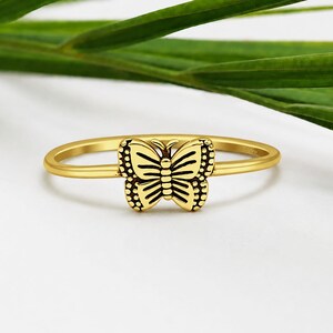 Könnte beinhalten: Ein goldfarbener Ring mit einem zentralen Schmetterlingsmotiv und schwarzen Gravurdetails auf den Flügeln. Der Ring ist dünn und kreisförmig und weist eine polierte metallische Oberfläche auf, die mit den dunklen Linien des Schmetterlings kontrastiert.