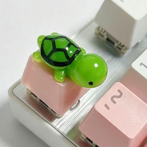 Puede incluir: Una tecla de teclado mecánico con una pequeña figura de tortuga verde brillante con un patrón de caparazón negro en la parte superior. La tecla es de color rosa claro y se encuentra entre otras teclas blancas y rosas con símbolos y números.
