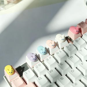 May include: A mechanical keyboard featuring white keys and a row of custom artisan keycaps shaped like small, colourful cats. The cat figures are painted in yellow, purple, light blue, peach, white, and pink, each with simple facial features.