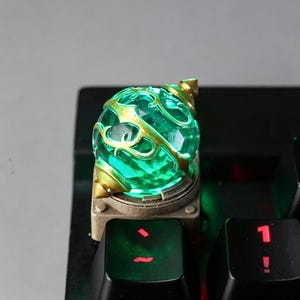 May include: A custom mechanical keyboard keycap featuring a translucent, glowing green orb encased in a gold-colored metal frame with intricate vine-like filigree details, mounted on a square base positioned on a black keyboard.