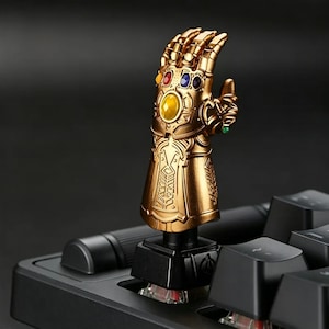 May include: A gold-colored mechanical keyboard keycap shaped like a gauntlet with intricate metallic detailing. The gauntlet features six embedded stones in yellow, red, blue, and green, mounted on a black square base that fits onto a keyboard switch.