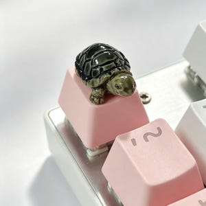 May include: A custom artisan keycap featuring a small, detailed, three-dimensional green and black turtle figurine mounted on top of a smooth, matte, light pink mechanical keyboard keycap, positioned next to a keycap marked with the number two.