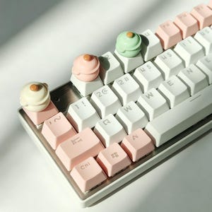 Puede incluir: Un teclado mecánico con teclas blancas y rosa claro. Tres teclas artesanales personalizadas con forma de ollas en miniatura con tapa están colocadas en la fila superior, en colores crema, rosa claro y verde menta respectivamente.