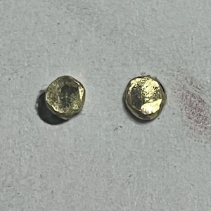 May include: Two small, gold-colored stud earrings. Each earring has a rounded, irregular shape with a textured surface. The earrings are displayed on a light gray surface.