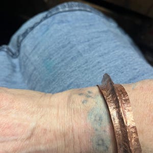 May include: A wide copper cuff bracelet featuring a highly textured, hammered surface with a prominent central ridge. The metal has a warm, reddish-brown metallic finish and a jagged, uneven edge profile that follows the organic, rippled form.