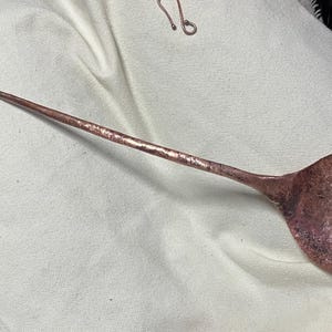May include: A handmade copper ladle with a long handle and a rounded bowl. The ladle has a hammered texture and a loop at the end of the handle. The copper has a reddish-brown color.