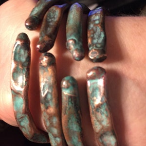 May include: A copper-toned bracelet with a hand-like design. The bracelet features six finger-shaped segments with a verdigris patina, giving it an aged appearance. The metal has a textured surface and a unique, artistic style.