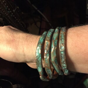 May include: Four copper bangles with a turquoise patina are stacked on a wrist. The bracelets have a hammered texture and a rustic, aged appearance. The copper is visible beneath the turquoise finish.