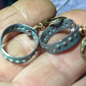 May include: Close-up of antique jewelry components. Two circular, oxidized metal rings with small, evenly spaced decorative elements. The rings are connected by gold-colored metal links. The overall aesthetic suggests vintage or handcrafted jewelry.