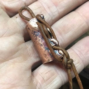 May include: A copper tube with a geometric pattern is wrapped with brown leather cord. The cord is tied in a knot and has a loop at the top.