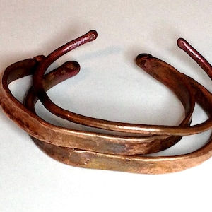 May include: Two open-ended cuff bracelets crafted from hammered copper. The metal features a warm, metallic reddish-brown tone with a textured, uneven surface and rounded, slightly thickened ends on each of the two circular bands.