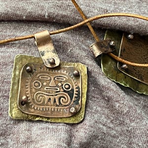 May include: Two handcrafted pendant necklaces. One pendant features a layered design with a copper face design on a brass base. The other pendant has a textured brass design. Both are on brown leather cords.