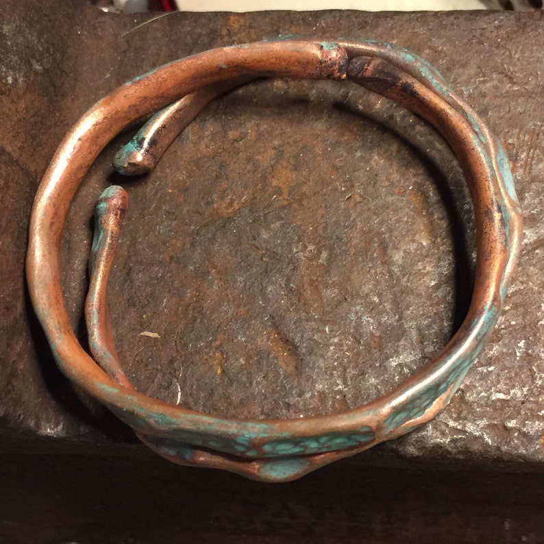 May include: A handcrafted copper bracelet with a unique, organic design. The bracelet features a textured surface with areas of turquoise-colored patina, adding visual interest. The open design allows for easy wear.