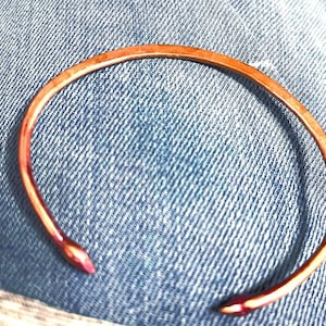 May include: A copper-colored open cuff bracelet with pointed ends. The bracelet has a hammered texture and is set against a blue denim background. The bracelet is a simple, yet elegant piece of jewelry.