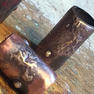 May include: Three metal cylinder beads with a rustic, hammered finish. The beads are a mix of copper and silver tones and have small silver accents.