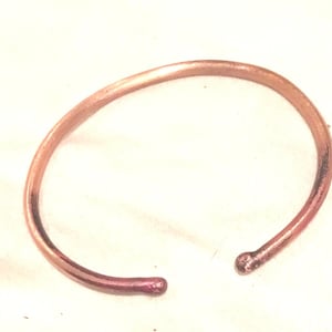 May include: A simple, thin, copper bracelet with a rounded, slightly hammered finish. The bracelet is a light copper color with a hint of pink.