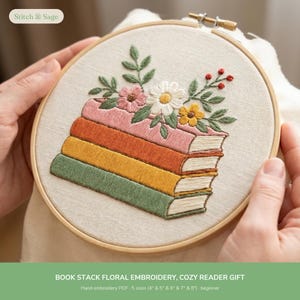 May include: A round wooden embroidery hoop containing a fabric panel with a hand-stitched design of a stack of three books in pink, orange, and green. A floral arrangement with white, pink, and yellow flowers and green leaves sits on top.