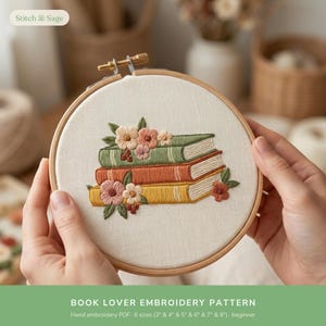 May include: A circular wooden embroidery hoop containing off-white fabric with a hand-stitched design of three stacked books in green, orange, and yellow. The books are decorated with embroidered pink, peach, and white flowers and green leaves.