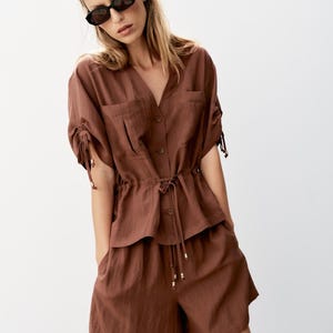 May include: A brown two-piece set consisting of a short-sleeved button-down shirt with a drawstring waist and matching shorts. The shirt features side-tie sleeves with metallic accents, a V-neckline, and two chest pockets. The shorts have a loose fit.