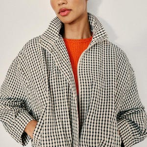 May include: A black and white gingham patterned jacket with a high collar and a front zipper closure, worn over a bright orange knit sweater. The jacket features long sleeves and side pockets, showcasing a classic checkered textile design.
