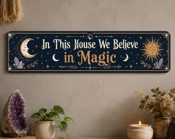 May include: A rectangular dark blue metal sign featuring the text In This House We Believe in Magic in white and gold lettering. The design includes a crescent moon with a face on the left, a sun with a face on the right, and purple crystals.