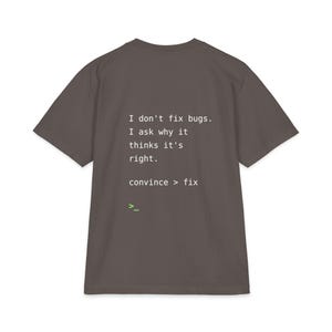 Può includere: Una t-shirt a maniche corte grigio scuro con testo bianco che recita: I don't fix bugs. I ask why it thinks it's right. convince > fix. Sotto il testo c'è un piccolo simbolo di prompt del terminale verde brillante composto da un segno maggiore e un trattino basso.