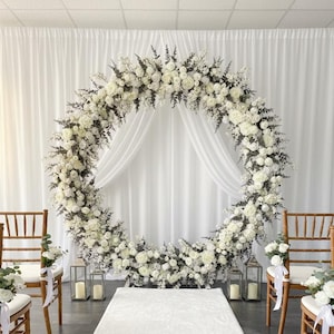 May include: A large circular wedding arch covered in white flowers and greenery, positioned in front of white draped curtains. Four wooden chairs with white floral accents line a white aisle runner, with metal lanterns placed near the arch base.