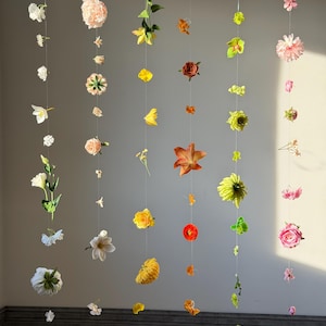 May include: Five vertical strings of artificial flowers in various colors including pink, yellow, orange, white, and green. Each string features a unique arrangement of blossoms, buds, and leaves suspended at different intervals along thin, clear lines.