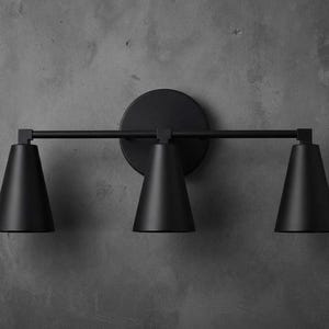 May include: A matte black wall-mounted light fitting featuring a horizontal bar attached to a circular backplate. The fitting includes three downward-facing conical shades arranged in a row, providing a uniform look for interior lighting.