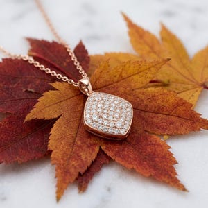 May include: A rose gold necklace featuring a square-shaped pendant with rounded corners, fully encrusted with small, clear, sparkling stones. The pendant hangs from a delicate rose gold chain, showcasing a polished metal finish.