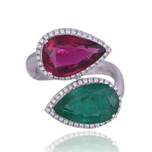 Puede incluir: Un anillo abierto con dos piedras preciosas en forma de pera, una de color rosa intenso y otra de color verde vibrante, rodeadas por un halo de pequeñas piedras blancas. La banda es de metal pulido en tono plateado.