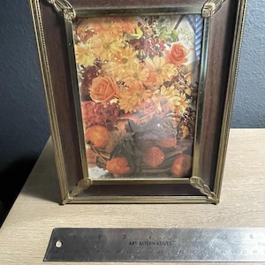 May include: A rectangular picture frame with a dark wood border and a gold-toned metal trim featuring decorative leaf-shaped corner accents. The frame holds a print of a floral arrangement and fruit. A metal ruler shows the frame is 8 inches wide.