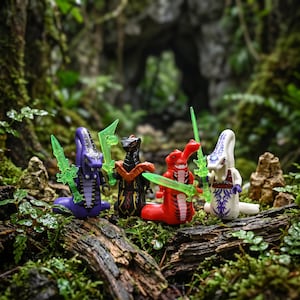 Könnte beinhalten: Vier Schlangen-Minifiguren stehen in einer Reihe. Die Figuren sind lila, schwarz, rot und weiß, haben jeweils markante Muster und halten durchscheinende grüne Plastikschwerter. Sie besitzen detaillierte Schlangenköpfe und Körper.