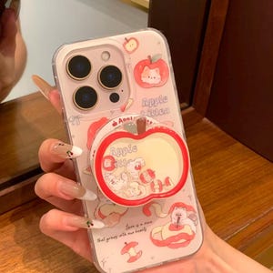 Puede incluir: Una funda transparente para smartphone con temática rosa y blanca, decorada con ilustraciones de gatitos y manzanas. Incluye un soporte para teléfono en forma de manzana roja con pequeños dijes flotantes y el texto Apple Kitten y Love is a vine that grows into our hearts.