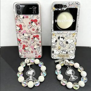 May include: Two Samsung Galaxy Z Flip5 phone cases with iridescent finishes. The left case features Snoopy in cowboy hats and boots. The right case shows various Snoopy poses. Both include matching wrist straps with iridescent beads and Snoopy charms.