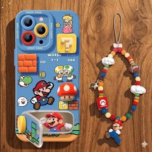 以下が含まれることがあります： スーパーマリオのキャラクターグラフィック、3Dキノコ、レンガブロック、マリオの絵が描かれた折りたたみ式ミラーが付いた水色のスマートフォンケース。「WORLD 1-1」や「TIME 666」という文字が記されています。お揃いのビーズストラップには、カラフルなビーズ、白い雲、フットボール、マリオのフィギュアがあしらわれています。