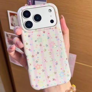 May include: A smartphone case featuring a light pink and white vertical striped pattern decorated with small, multicolored holographic stars. The case has a white camera cutout area with three circular lens openings and a flash.