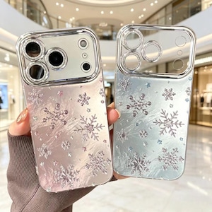 May include: Two smartphone cases featuring a metallic finish and a raised snowflake pattern. The left case has a pinkish hue, while the right case is light blue. Both cases include integrated camera lens protectors with a silver, crystal-like border.